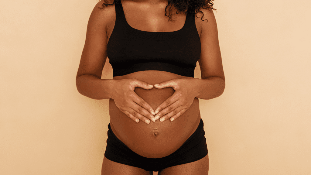 Mulher negra grávida  fazendo sinal de coração na barriga. 