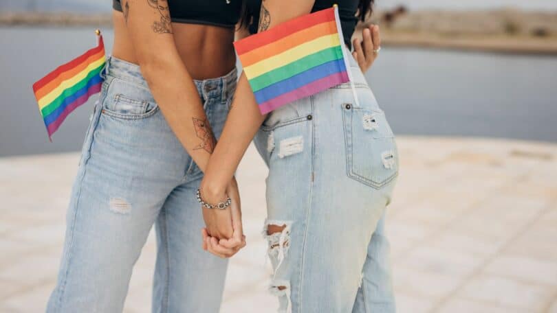 Duas mulheres segurando as mãos e a bandeira LGBTQIA+