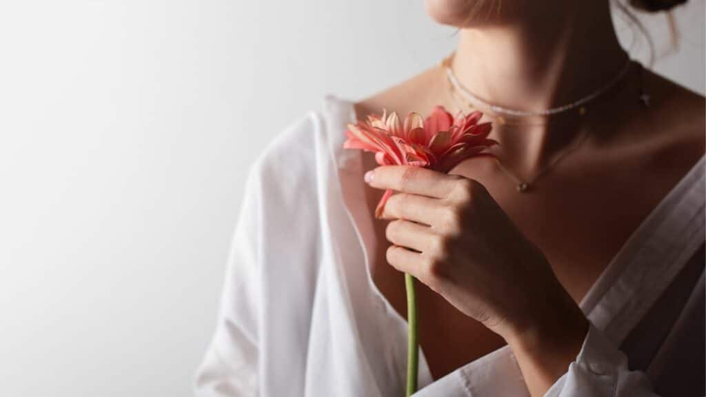Imagem só do pescoço pra baixo de uma mulher de camisa branca, que está segurando uma flor vermelha