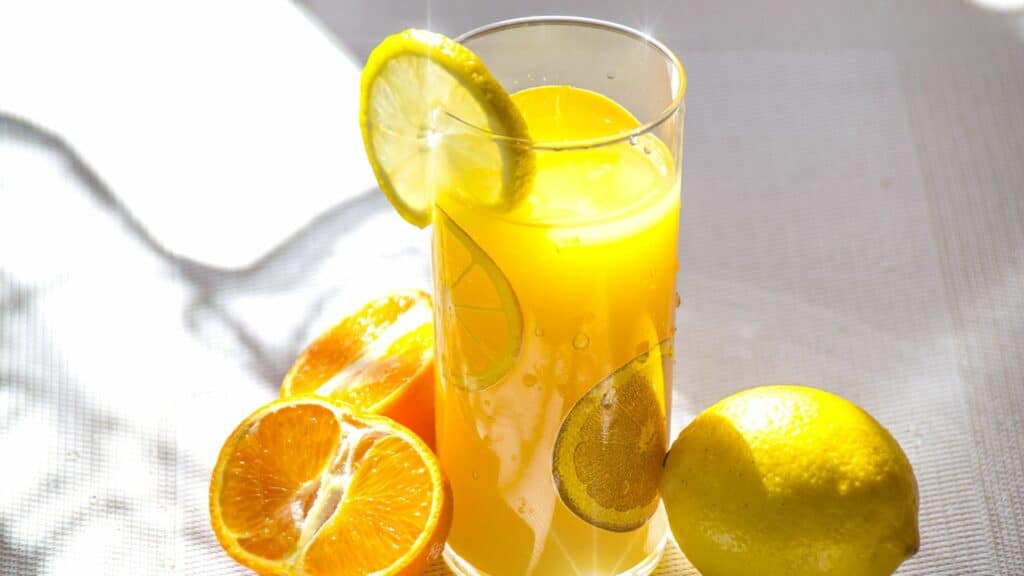 Imagem de um suco de laranja, com limões dentro do suco, e fora também, na mesa.
