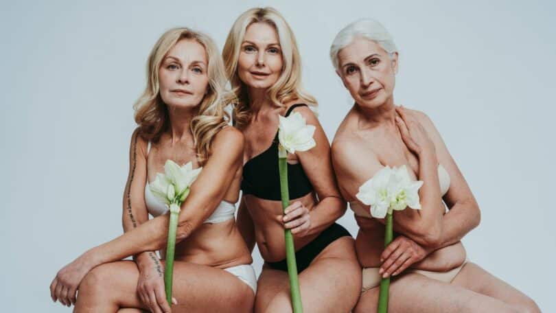 Imagem de três mulheres de meia idade, semi-nuas cada uma segurando uma flor branca.