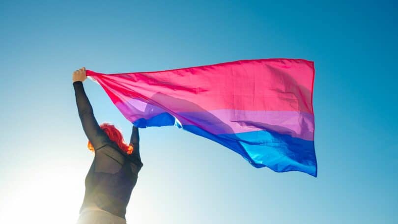 Imagem de uma moça segurando a bandeira da bissexualidade e o céu azul na paisagem