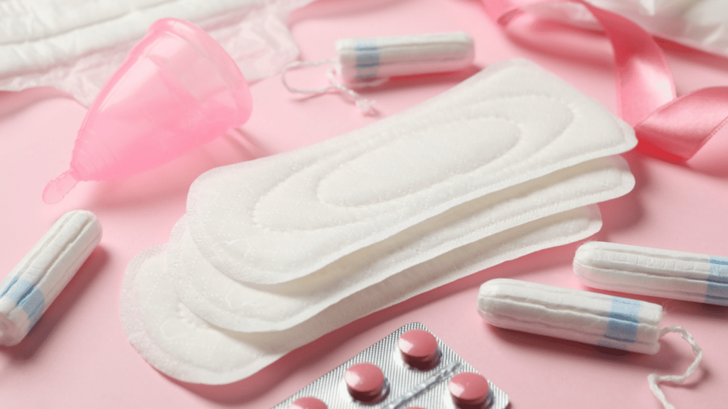 Vários absorventes, obs, coletor menstrual e remédios dispostos em um fundo rosa