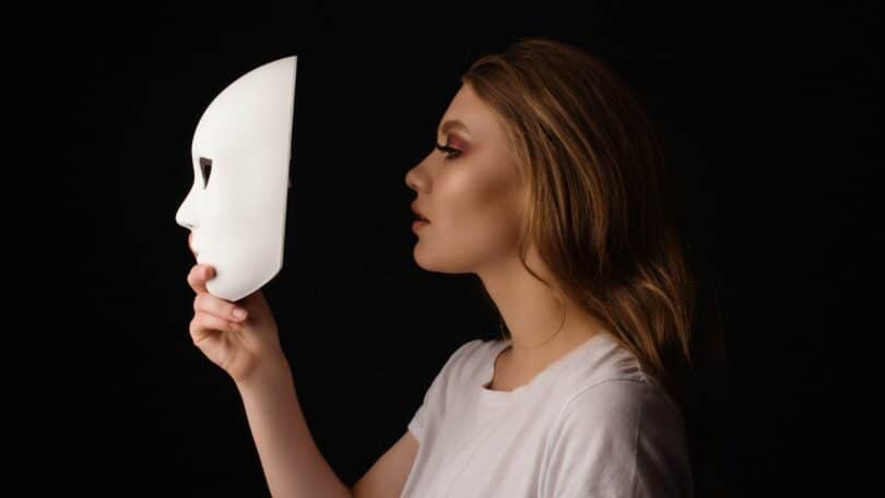 Imagem de uma mulher segurando uma máscara branca na frente do rosto, como se estivesse se escondendo