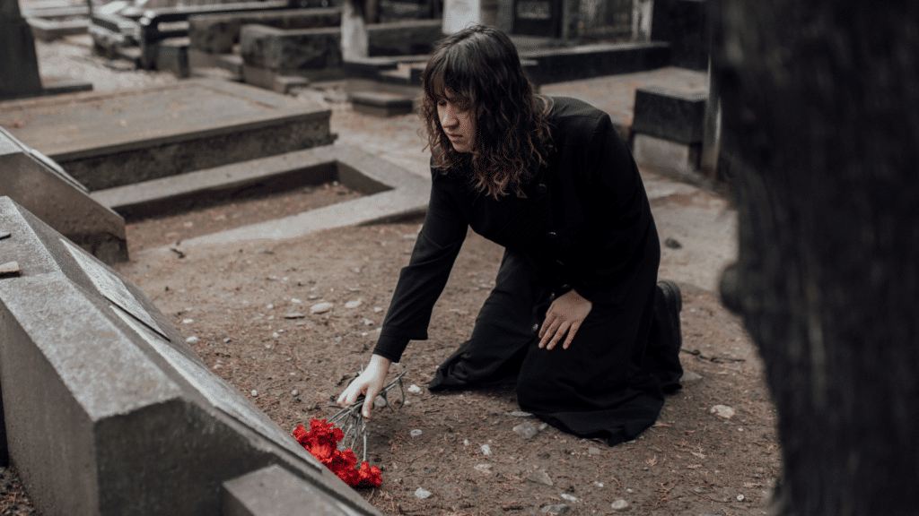 Mulher vestindo casaco preto, ajoelhado em frente à uma cova, entregando flores para um falecido. 