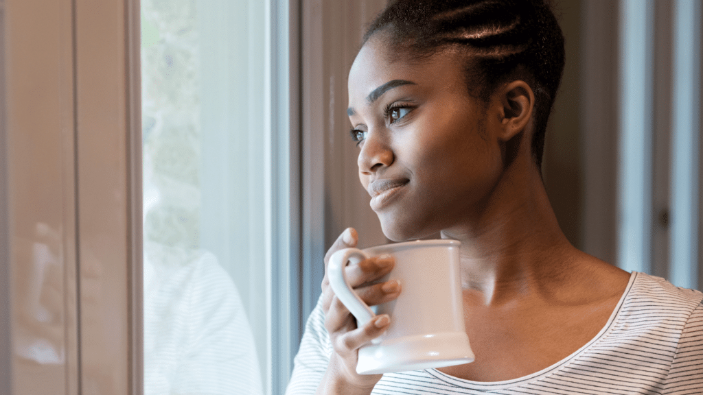 Mulher negra e jovem olhando de forma reflexiva por uma janela enquantos segura uma xícara de café. 