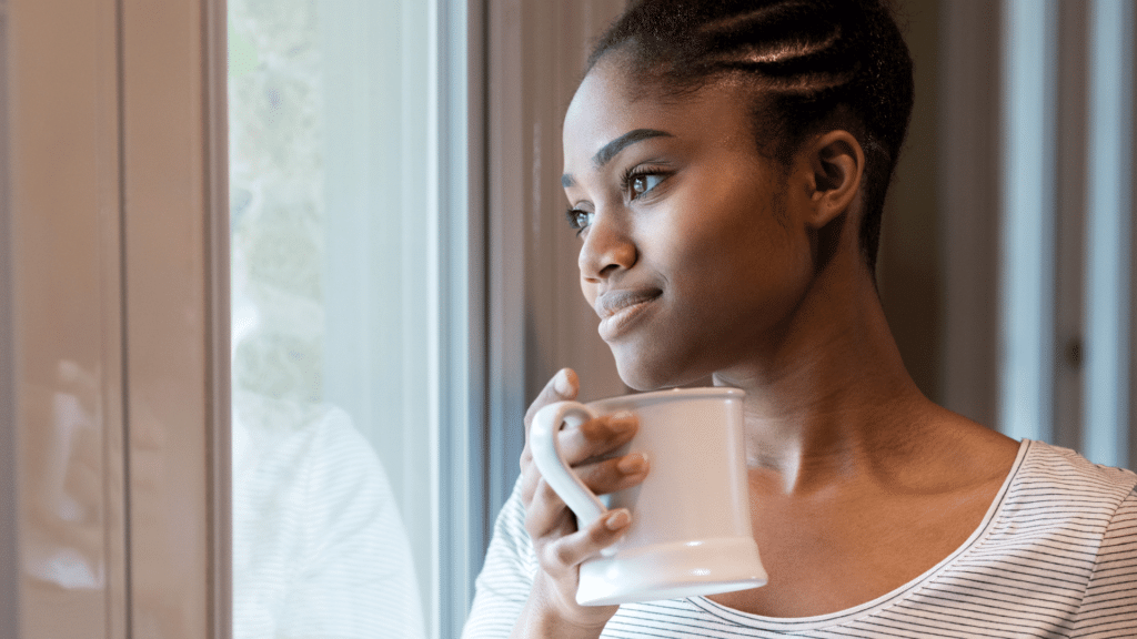 Mulher negra e jovem olhando de forma reflexiva por uma janela enquantos segura uma xícara de café.