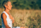 Mulher meditando no meio da natureza, com a mão sobre o peito. Representação de positividade e resiliência.