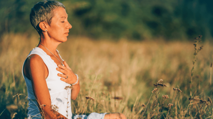 Mulher meditando no meio da natureza, com a mão sobre o peito. Representação de positividade e resiliência.