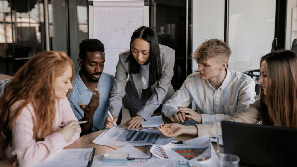 Funcionários reunidos em uma mesa, atentos ao um projeto empresarial que sua chefe os aponta.