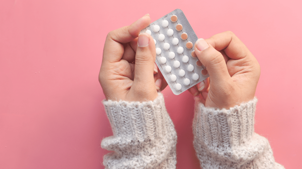 Mãos brancas de uma mulher segurando pílulas anticoncepcionais, em fundo rosa.
