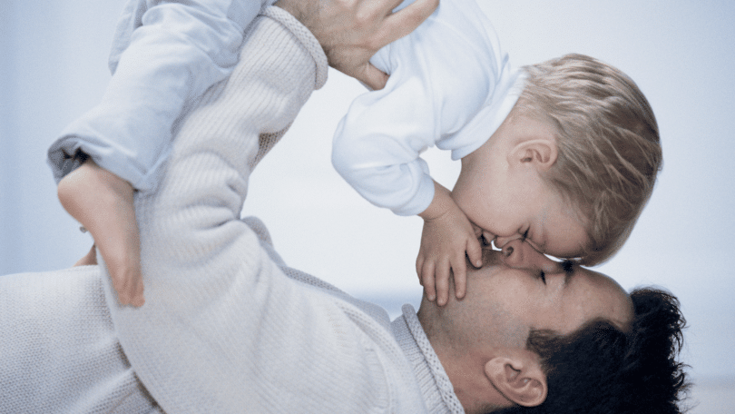 Pai deitado no chão de costas, segurando seu filho ao alto enquanto o beija.