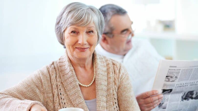 senhora sorrindo com uma xícara na mão e atrás dela um senhor lendo jornal