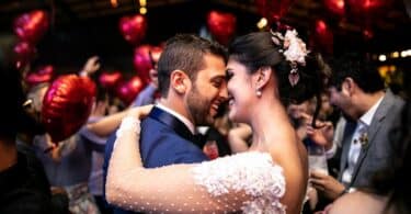 Casal de noivos abraçados e sorrindo e a festa acontecendo atrás deles