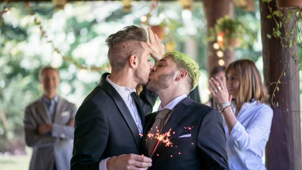 Casal gay se beijando com pavil de luz na mão, em sua festa de casamento e pessoas aplaudindo atrás