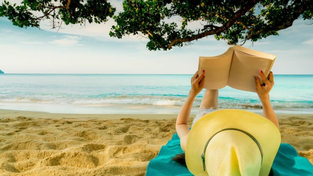 Mulher deitada na areia da praia, debaixo de uma árvore e de frente para o mar lendo um livro