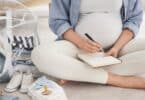 Mulher grávida fazendo uma lista do que levar na bolsa maternidade, ao lado dela, itens do bebê