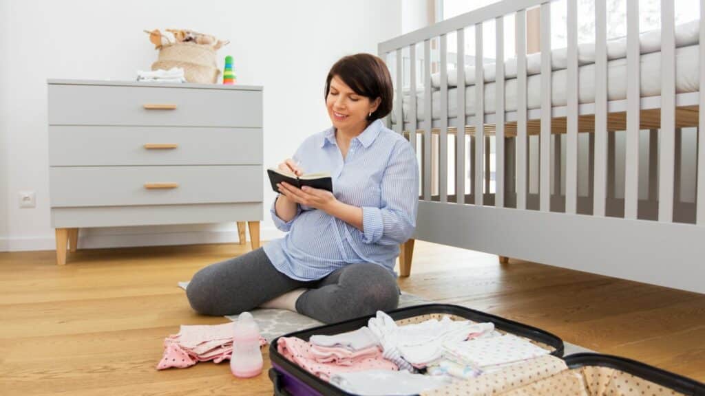 Mulher no quarto do seu bebê, montando uma bolsa maternidade para ele, com um caderno para o checklist e sorrindo