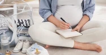 Mulher grávida fazendo uma lista do que levar na bolsa maternidade, ao lado dela, itens do bebê