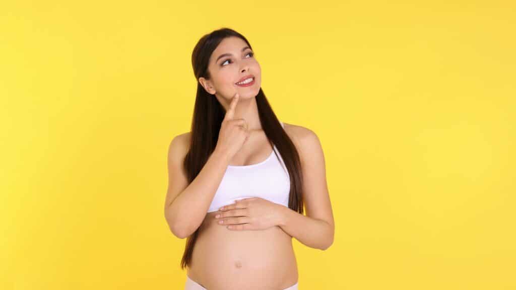 Mulher grávida, com a mão no queixo, como se estivesse com dúvida, mas está sorrindo