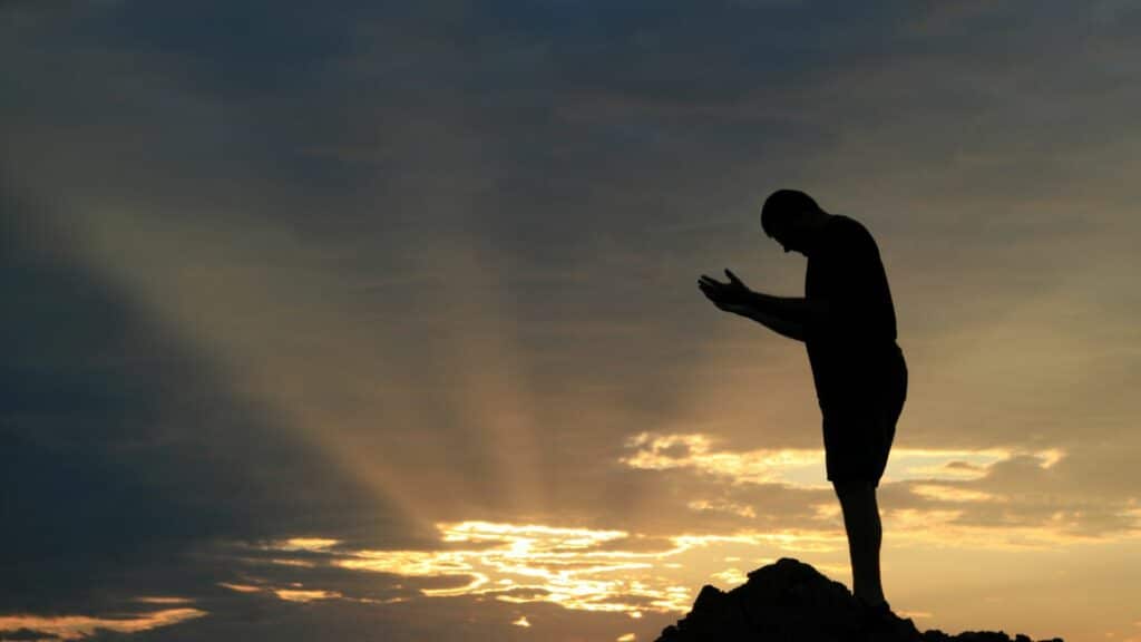 Homem no topo de uma montanha ao pôr-do-sol com a cabeça baixa e as mãos em posição de "receber"