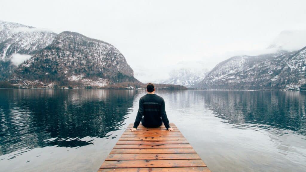 Homem sentado em uma pequena ponte de frente para o lago, em meio as montanhas com neve