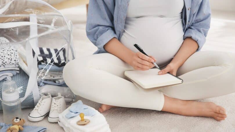 Mulher grávida fazendo uma lista do que levar na bolsa maternidade, ao lado dela, itens do bebê