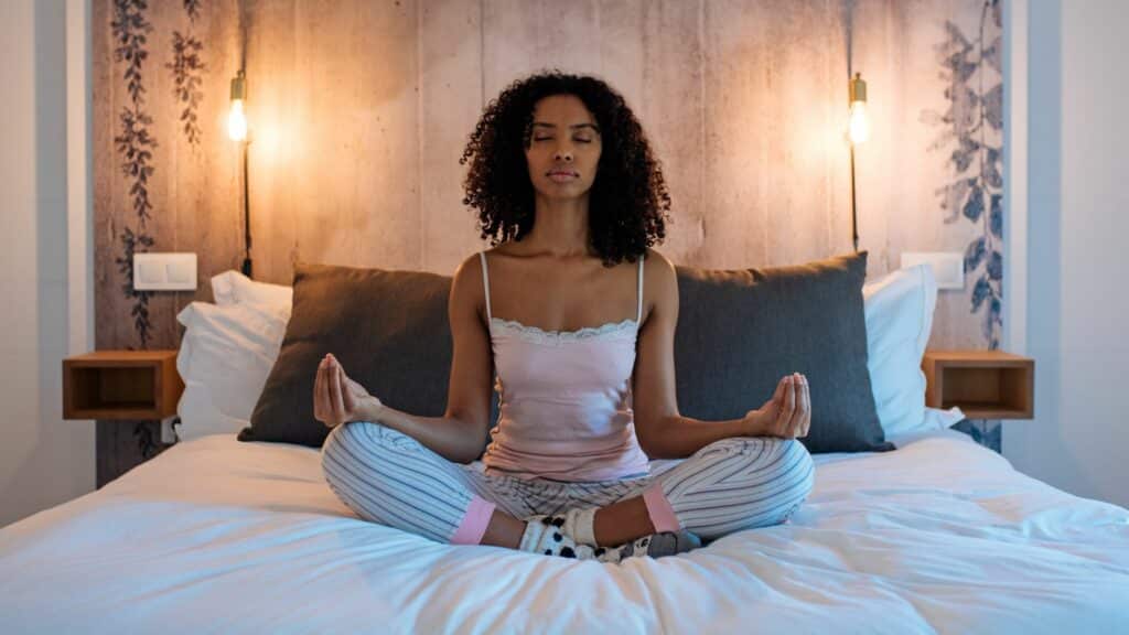 Mulher negra sentada na cama, com pernas cruzadas, enquanto medita.