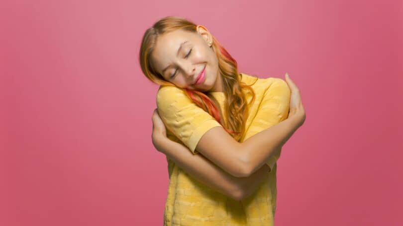 Mulher de amarelo, abraçando a si mesma em um fundo rosa