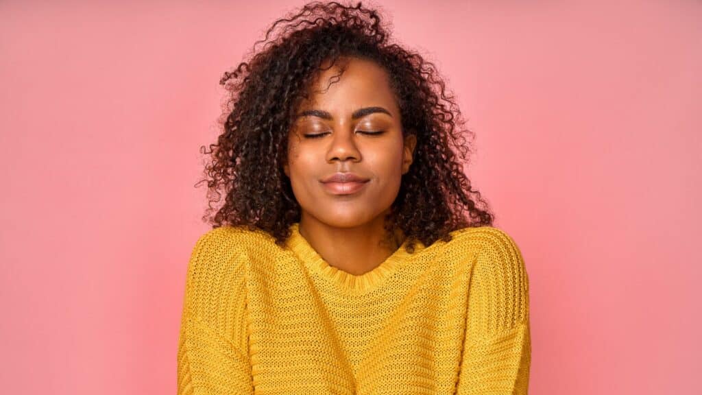 Mulher negra com roupa amarela, de olhos fechados e com um sorriso pacífico no rosto, em um fundo rosa