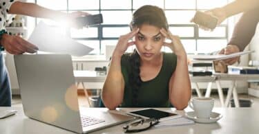 Mulher na frente de seu laptop, com algumas pessoas ao seu redor solicitando várias coisas ao mesmo tempo, e o seu semblante é de estresse