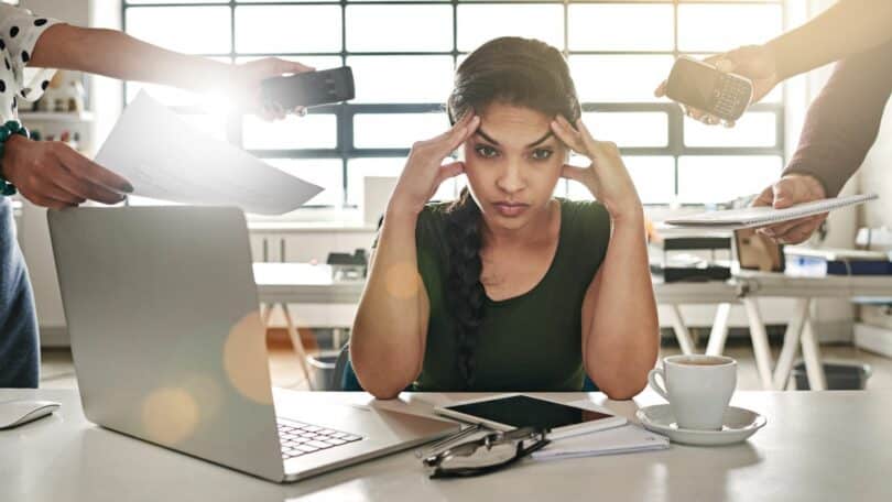 Mulher na frente de seu laptop, com algumas pessoas ao seu redor solicitando várias coisas ao mesmo tempo, e o seu semblante é de estresse