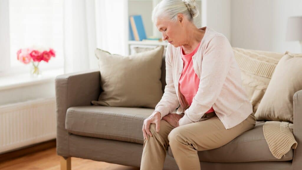 Uma senhora sentada no sofá com as mãos nas pernas como se estivesse sentindo dor