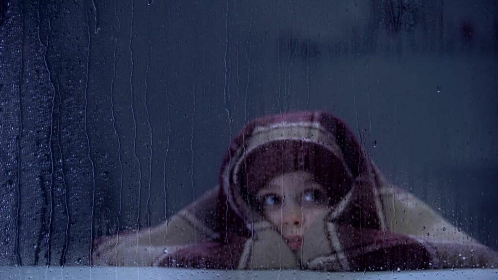 Criança em frente à janela, enrolada em cobertor e assustada com a chuva de tempestades.