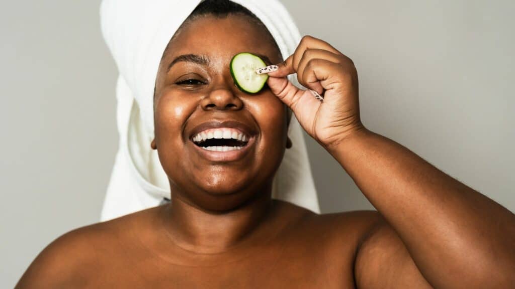 Mulher negra sorrindo para câmera, segurando uma fatia de pepino no olho. Conceito de autocuidado e autoestima.