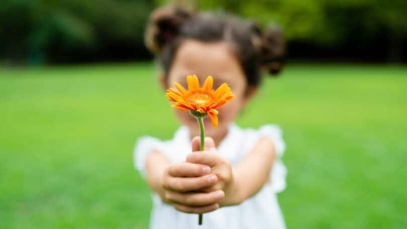 Garotinha segurando uma flor laranja nas mãos. Ela está sobre um ambiente verde, com árvores e grama. O fundo está embaçado (efeito da foto).