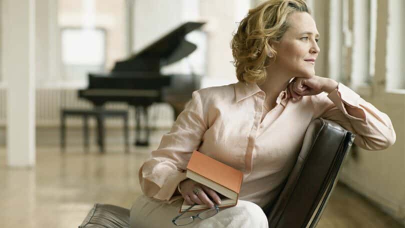 Mulher olhando pela janela, sentada em uma poltrona e com um livro semi-aberto na mão.
