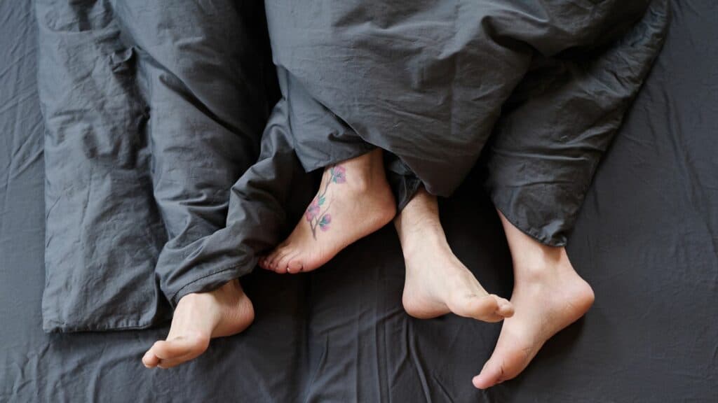 Pés de casal na cama, em lençóis cinzas.