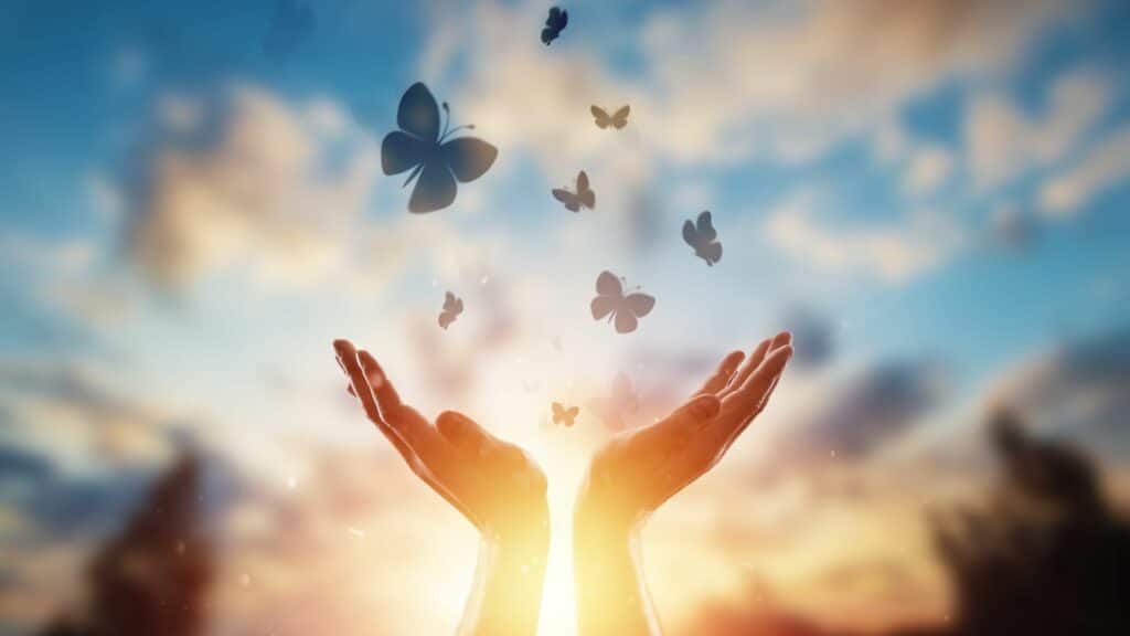 Imagem de um céu com núvens e uma mão aberta em sinal de gratidão e algumas borboletas voando no céu.