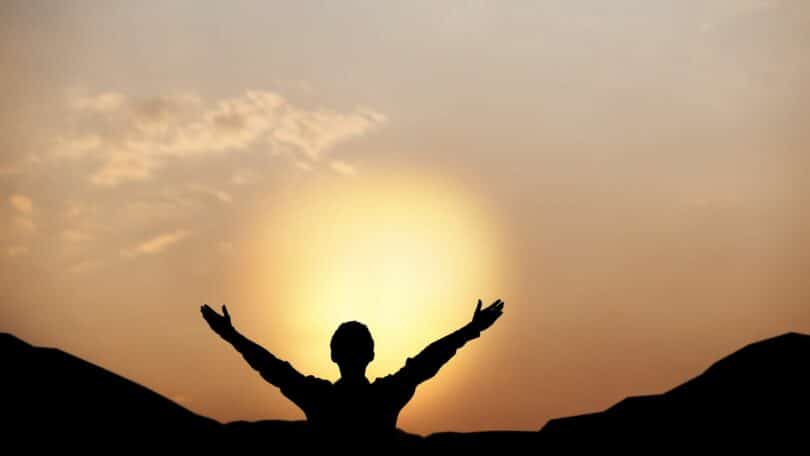 Imagem do pôr do sol e em destaque a silhueta de um homem olhando para o so de braços abertos.