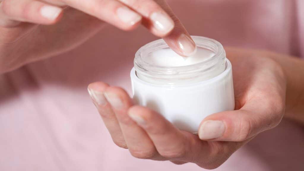 Mãos femininas segurando creme nas mãos. O creme está aberto e ela está com dedo dentro do pote.