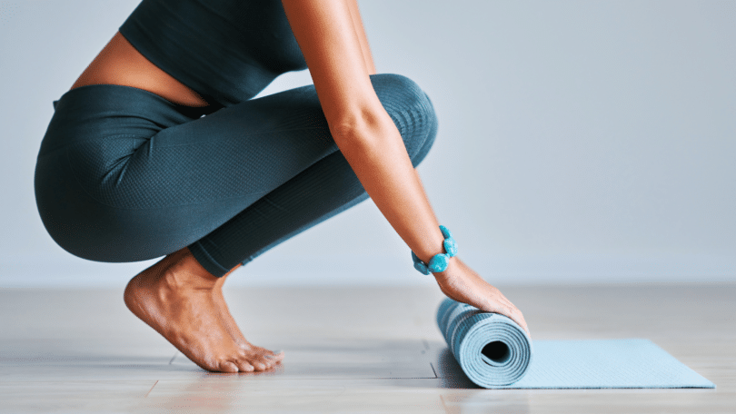 Mulher agachada no chão, abrindo tapete azul de yoga.