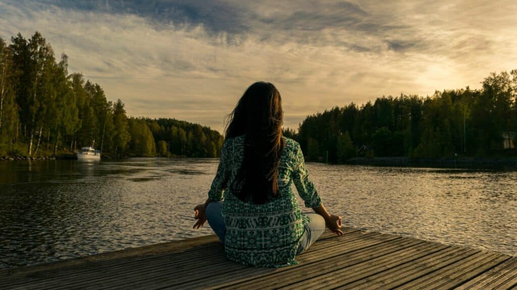 Imagem de fundo de um rio e em destaque uma mulher de cabelos longos, sentada sobre uma ponte meditando.