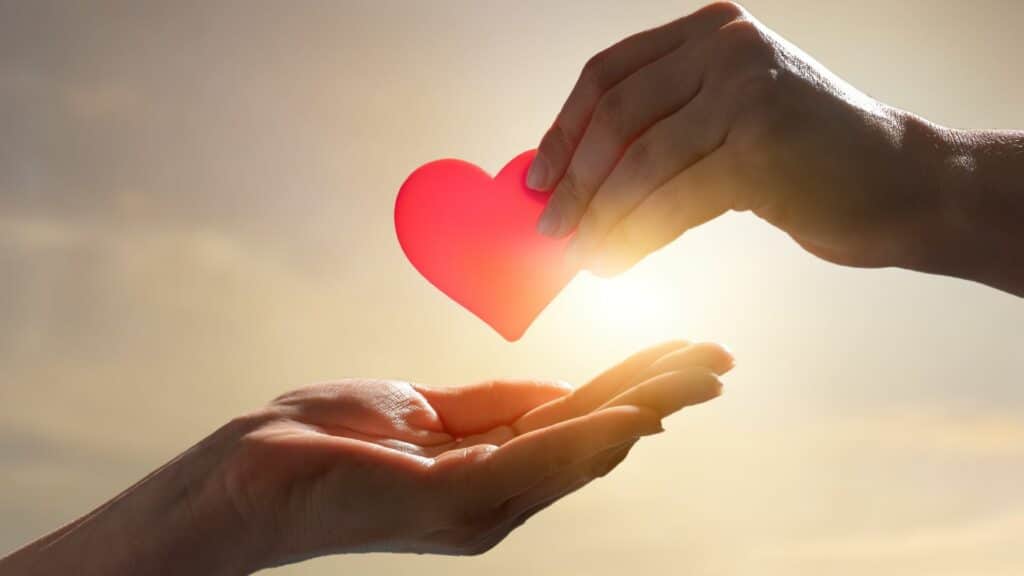 Imagem com um lindo por do sol ao fundo e em destaque duas mãos, sendo que uma delas segura um pequeno coração vermelho.
