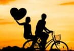 Imagem de um céu alaranjado e em destaque a silhueta de um casal (homem e mulher) sentados sobre uma bicicleta. A mulher está na garupa e segura um coração ao alto.