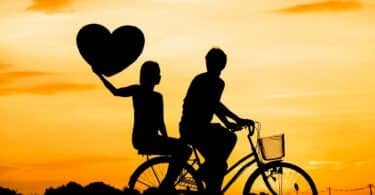 Imagem de um céu alaranjado e em destaque a silhueta de um casal (homem e mulher) sentados sobre uma bicicleta. A mulher está na garupa e segura um coração ao alto.