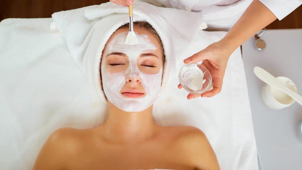 Imagem de uma mulher deitada sobre uma maca tratando a pele do rosto. Ela usa uma máscara facial na cor branca.