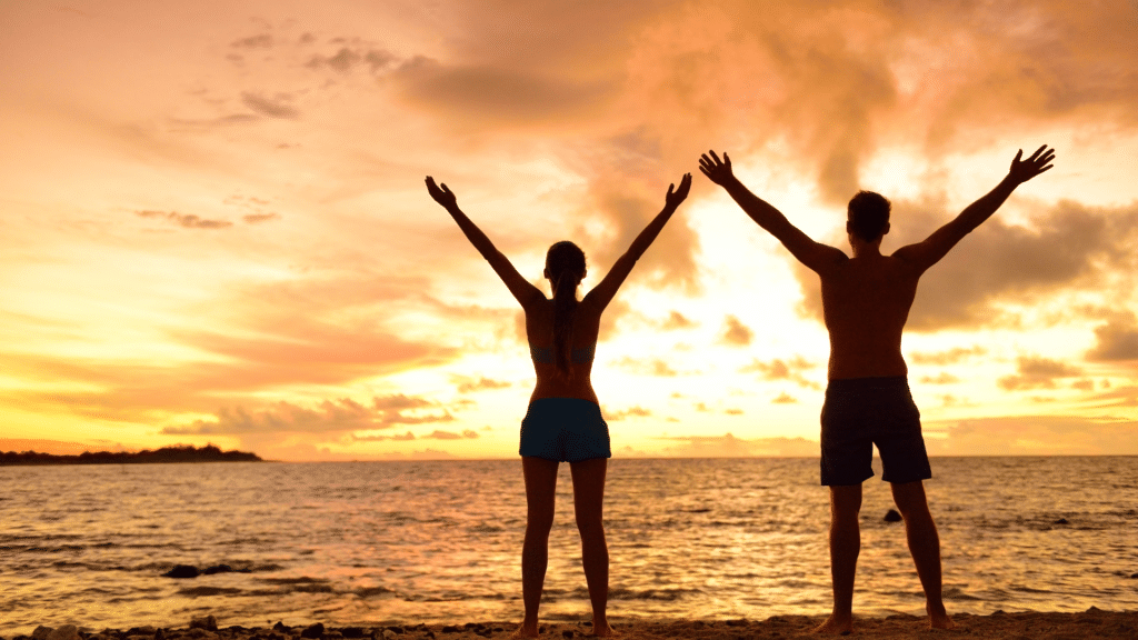 Imagem de fundo de um lindo pô do sol na praia. Em destaque um casal contemplando a beleza e a liberdade.