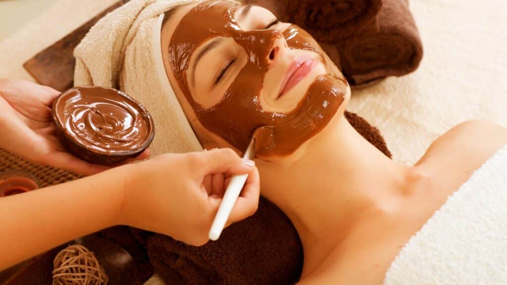 Imagem de uma mulher deitada sobre uma maca, com uma toalha no cabelo. Ela está usando uma máscara de chocolate na pele do rosto.