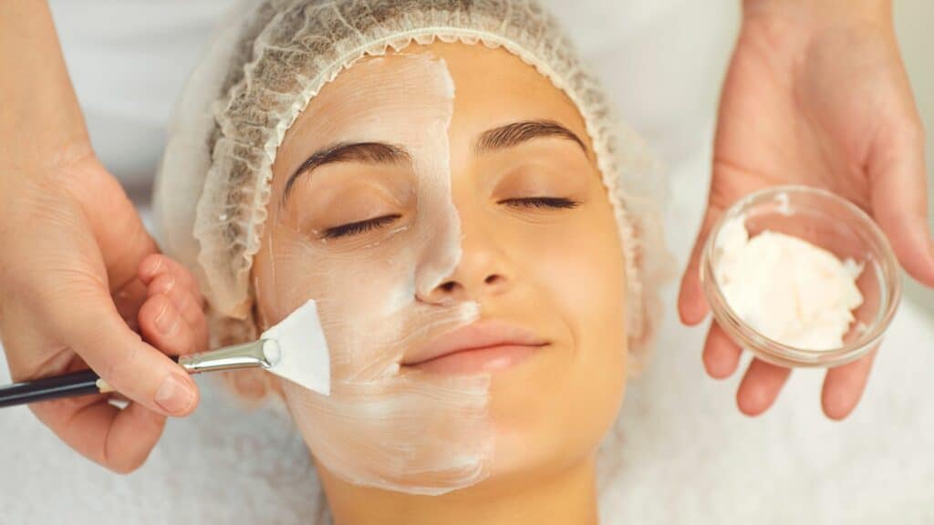 Mãos de especialista em cosmetologia aplicando máscara facial branca com pincel, deixando a pele hidratada.